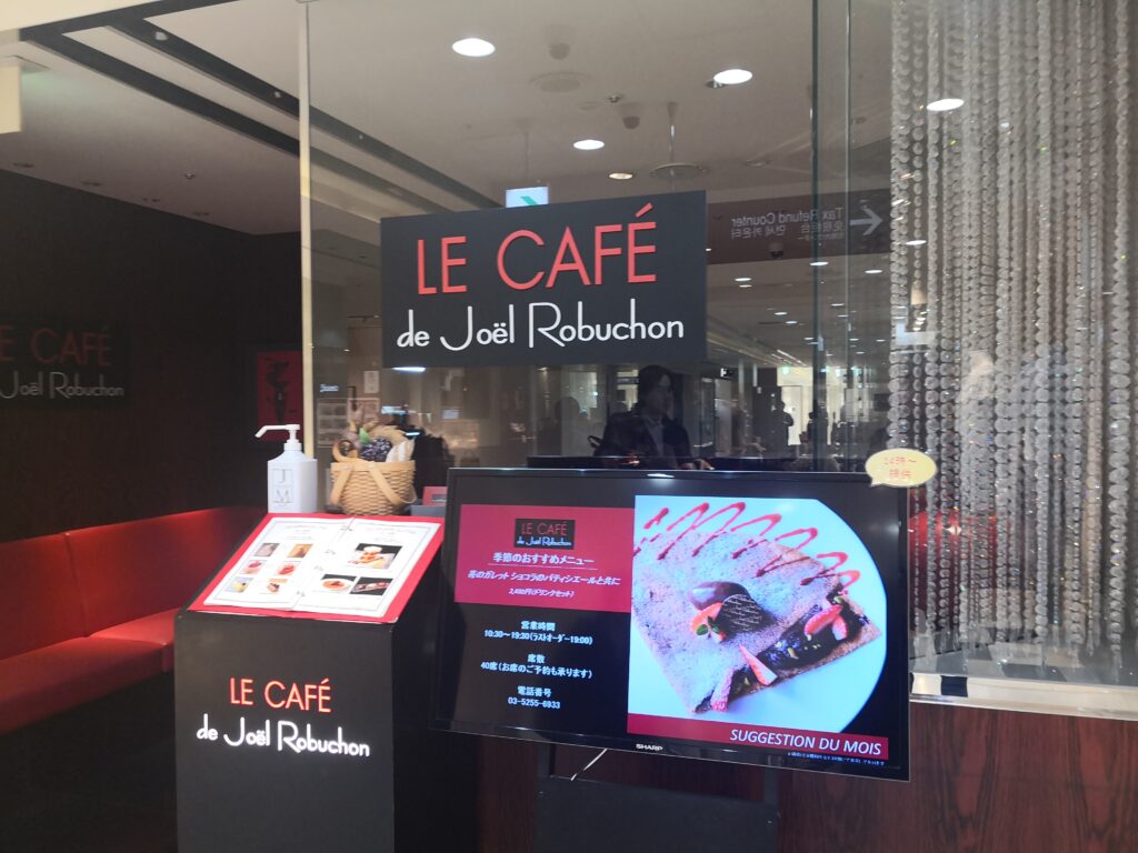 （店舗外観）
ル カフェ ドゥ ジョエル・ロブションの店舗外観。ガラス張りのエントランスに、黒地へ赤の「LE CAFÉ de Joël Robuchon」のロゴサイン。手前のモニターには季節のおすすめメニューが表示されている
