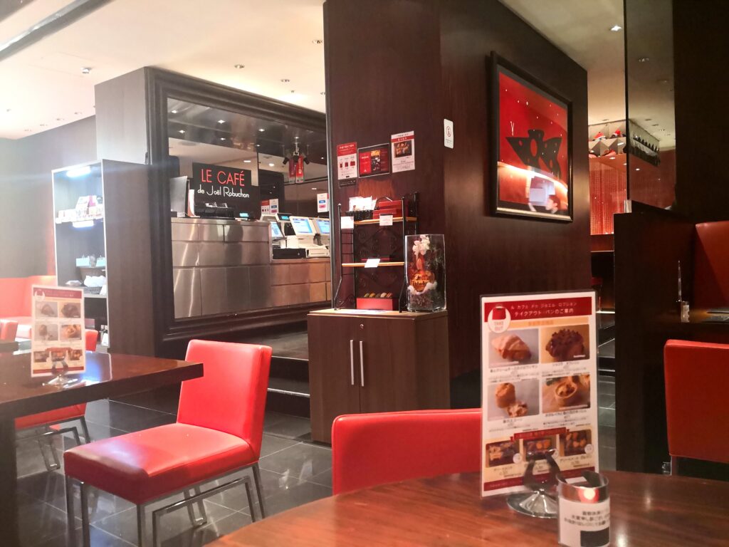 （店内の雰囲気）
赤いレザーの椅子と黒を基調としたル カフェ ドゥ ジョエル・ロブションの店内。壁にはロブションのアートが飾られ、テイクアウトメニューのスタンドが置かれている