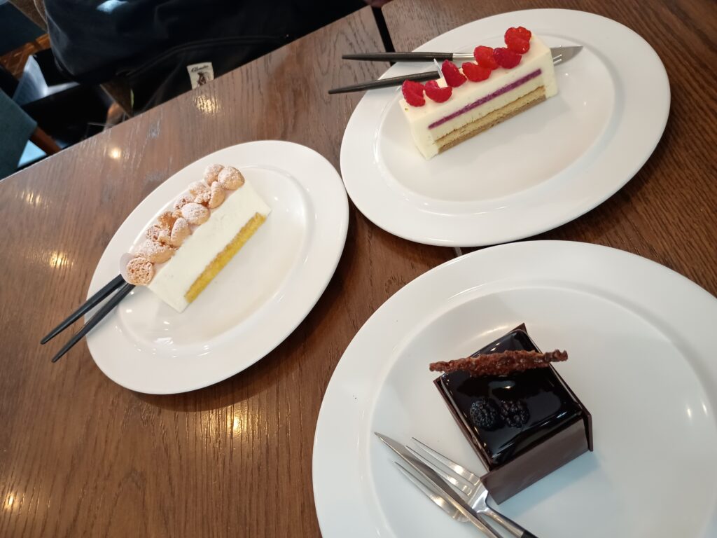 パリセヴェイユのケーキ3種。木のテーブルに白い皿が3枚。手前右がツヤツヤのチョコレートコーティングにブラックベリーをのせた「ル・シュープレーム」、左が粉糖をまぶした小さなシュー生地を並べた「コム ニューヨーカー」、奥がフランボワーズをのせた白いムースに紫の層が見える「ジベルニー」。
