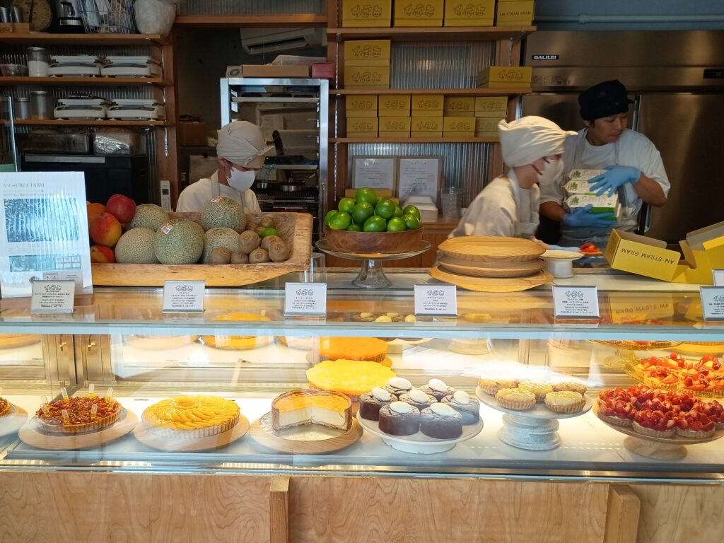 店内・ショーケース全景
アムストラムグラムのショーケースと作業スペース全景。カウンター上にはメロンやライムなど旬のフルーツが並び、マスク姿のスタッフ2名が仕込み作業中。ガラスケース内には色とりどりのタルトが並ぶ。棚にはブランドの黄色い箱が整然と積まれている
