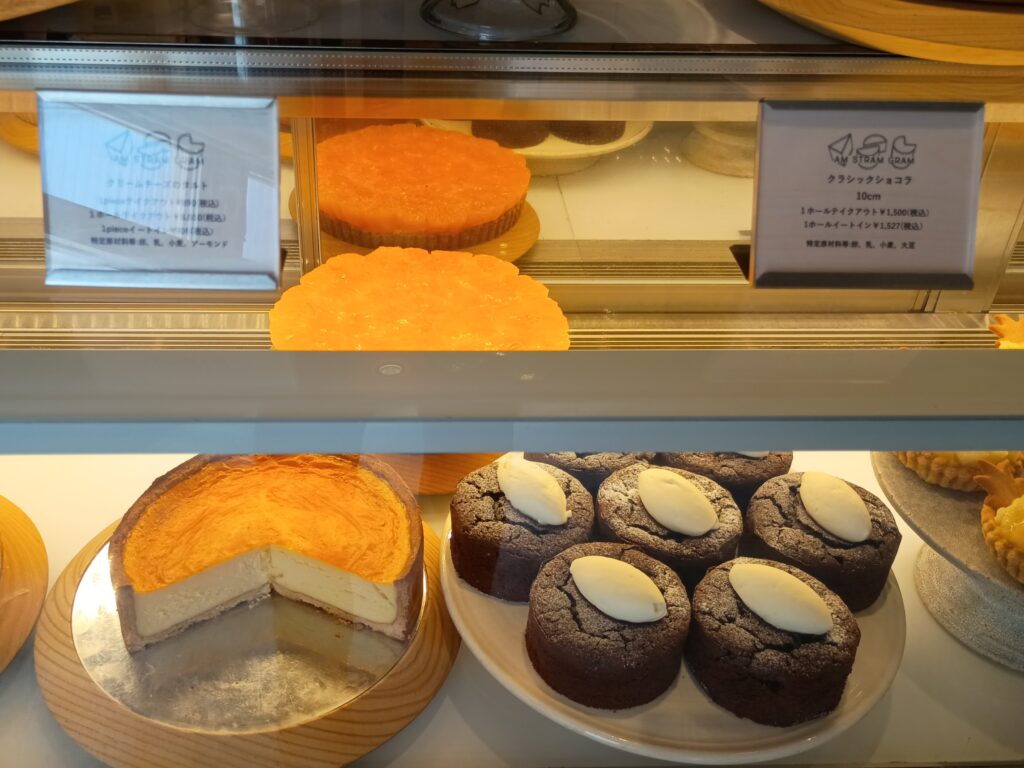 ショーケースに並ぶクリームチーズのタルト（左、こんがりした焼き色のホール）とクラシックショコラ（右、白いクリームをのせた小ぶりなショコラが4個）。上段にはオレンジ色のタルトが見える
