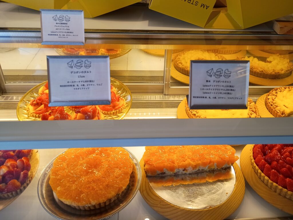 ショーケースに並ぶデコポンのタルト2種。手前左は丸いホールタイプ、右は角型でカット断面が見えている。デコポンの鮮やかなオレンジ色が映え、上段には各商品のPOPカードが立てられている
