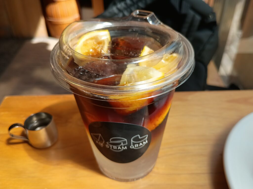  コーヒートニック＆シトラス・ドリンク。アムストラムグラムのロゴ入りクリアカップに注がれた期間限定ドリンク「コーヒートニック＆シトラス」。深いブラウンのコーヒートニックに黄金柑のスライスが浮かび、クリアなカップから鮮やかな色合いが透けて見える