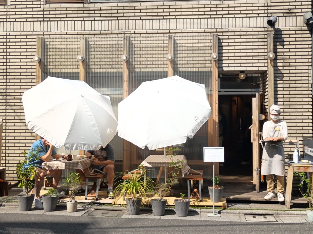  店舗外観・表側（10165 / 141337503）
アムストラムグラム恵比寿店の外観。薄いレンガ調の壁面と木製フレームが特徴的で、白いパラソルのテラス席が2組並ぶ。入口脇にはグレーのエプロン姿のスタッフが立ち、鉢植えが並んだ開放的な雰囲気
