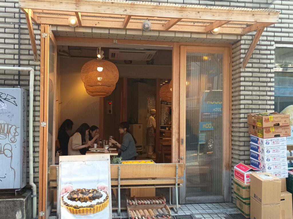  店内・裏側（10166 / 141807826）
アムストラムグラムの店内を通り側から見た様子。ラタン素材の大きな吊り照明が温かみのある光を放ち、女性客がテーブルで談笑している。右手には隣接する青果店が見え、代官山らしい生活感のある一角
