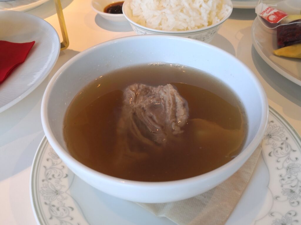 （バクテー）
バクテー（肉骨茶）。澄んだ茶色いスープに骨付き肉が入った白い器、奥に白米