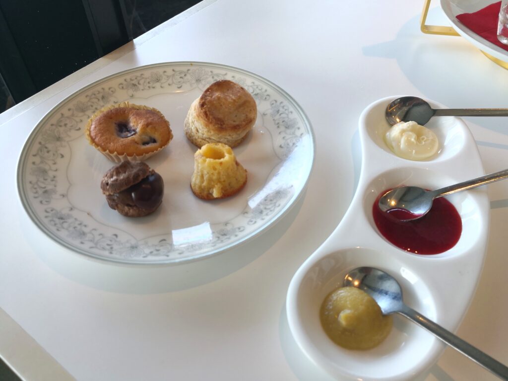 スコーン＆ジャム）
取り分けたスコーンやマフィンの皿と、クロテッドクリーム・チェリージャム・カヤジャムが並ぶ3連の小皿